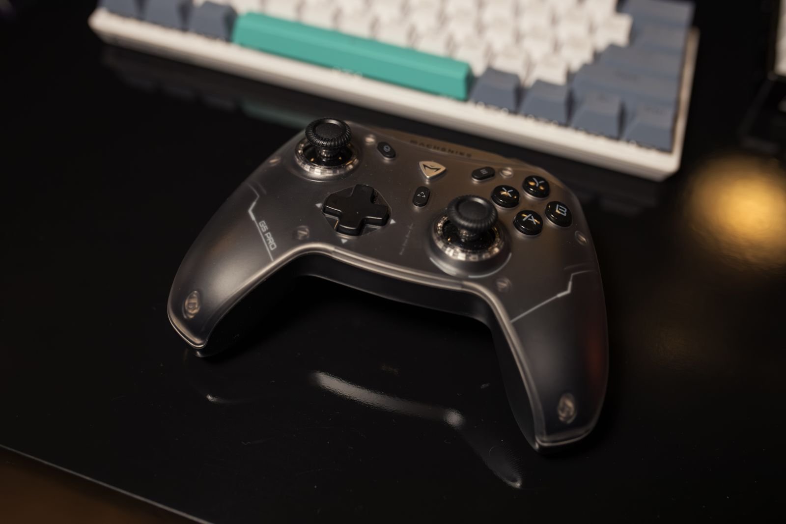 Black game controller resting on a dark desk in front of a white and teal keyboard.