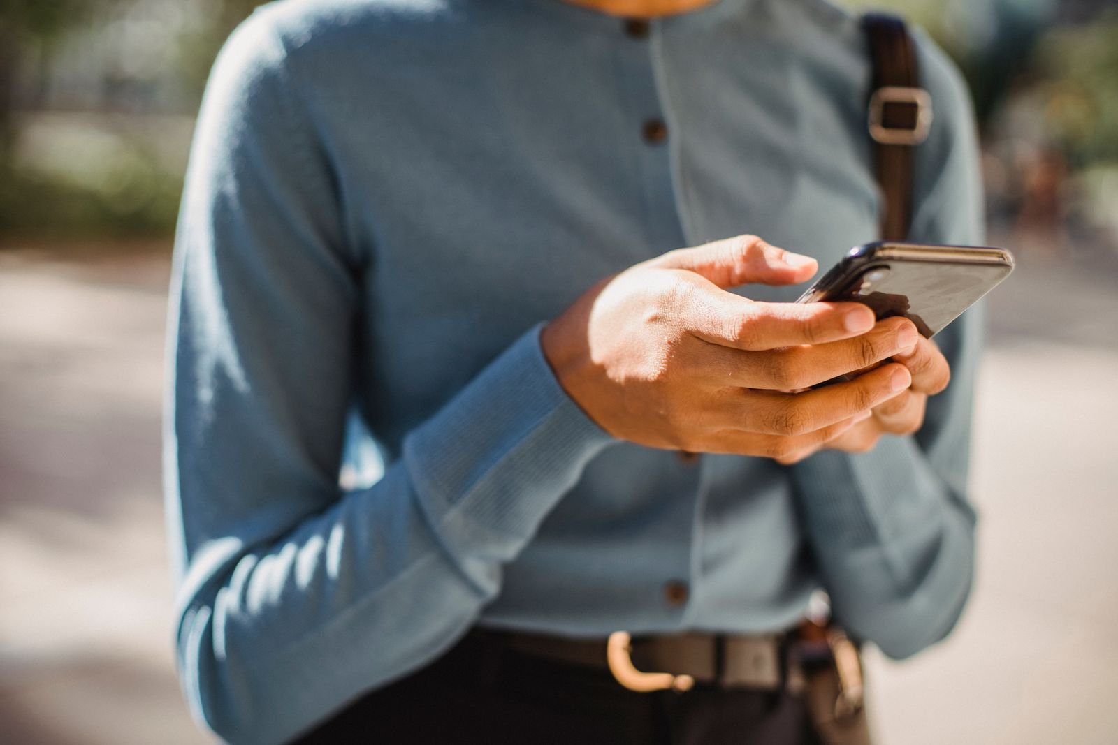 Person in a light blue top using a smartphone outdoors, with their hands and phone in focus.